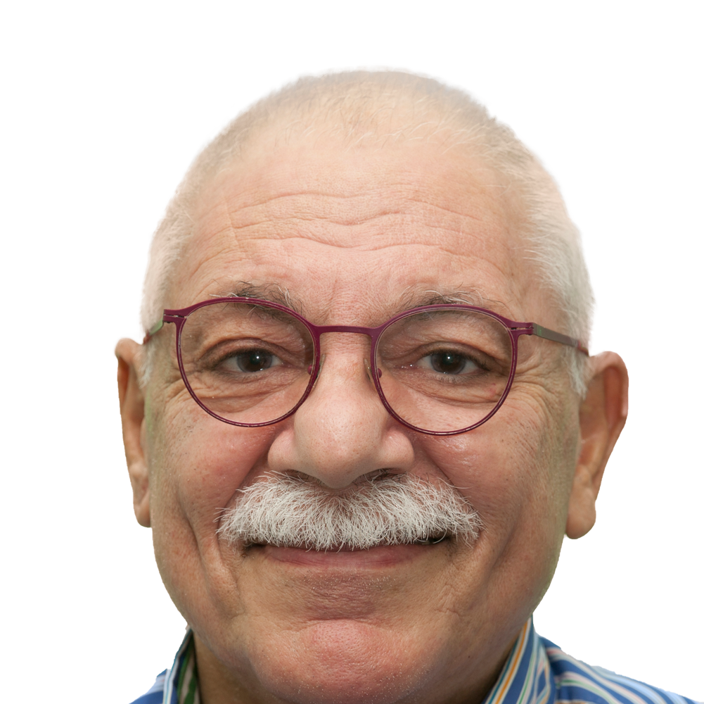 An older man half smiles at the camera. He has a thick white mustache and short white hair. He has hazel coloured eyes, and wears oval shaped glasses that have a dark red frame. He is wearing a shirt with white and blue stripes.