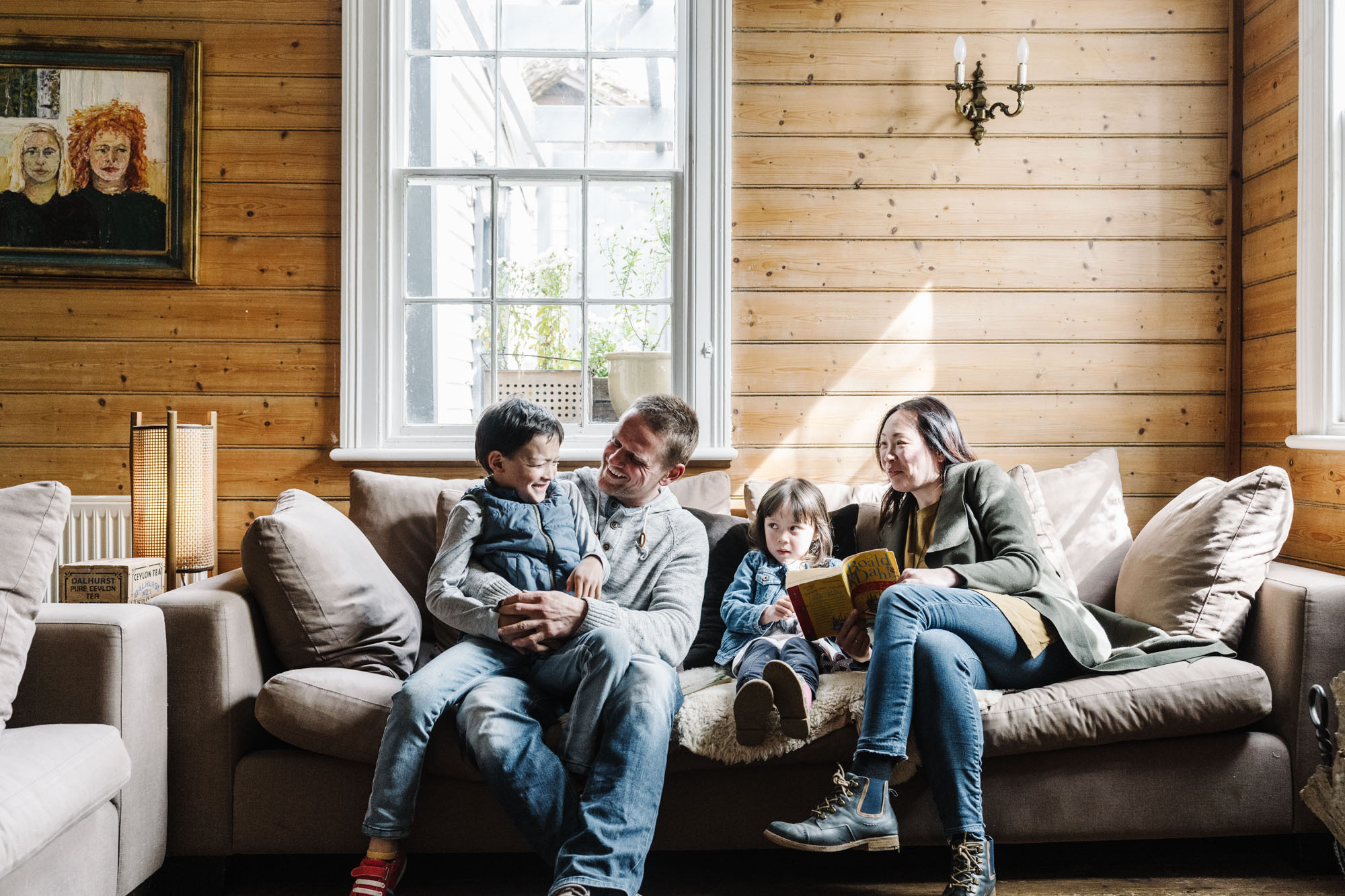 A family of four sits on a couch inside a cabin. The father has his son, aged around five, sitting on his knee. The mother is reading a book with their young daughter who is a toddler. The family looks happy and are laughing together.