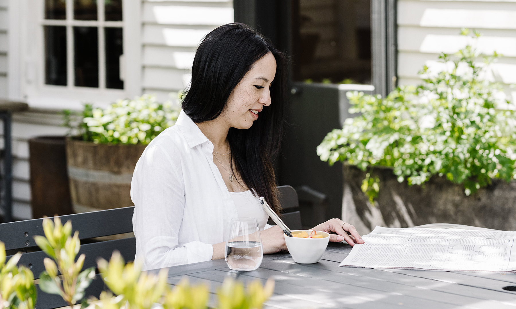 A woman sits outside, eating breakfast and reading the newspaper. She is surrounded by green plants.