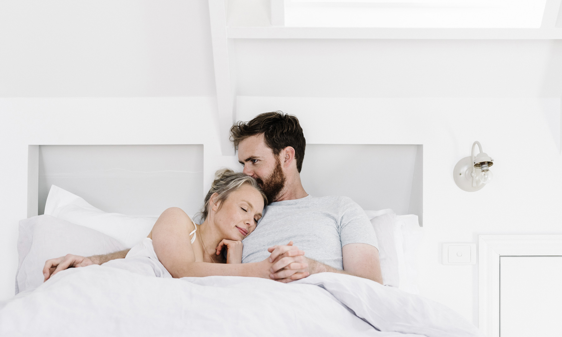 A man and woman cuddle in bed next to each other. The woman rests her head on the man's chest, and he kisses her head.